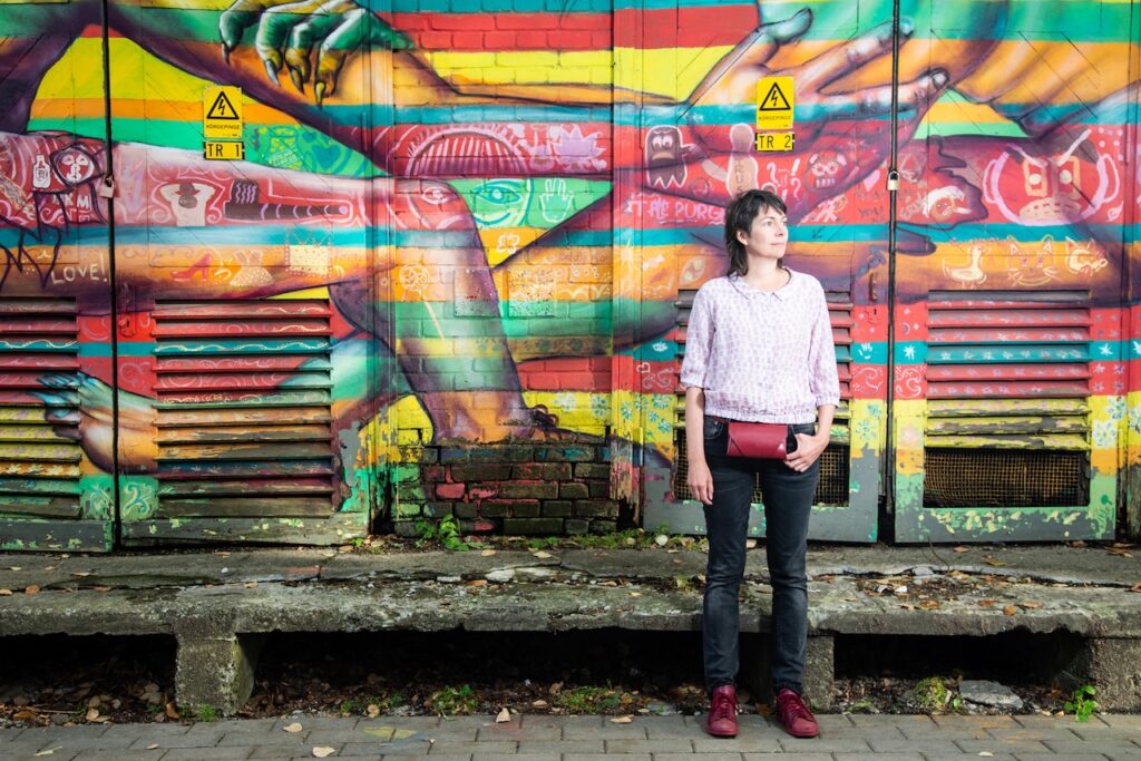 Naine seisab erksavärvilise graffitiga kaetud seina ees. Tal on mullet ja ta kannab roosakirjut toppi, kulunud musti teksapükse, punaseid sneakerseid ja väikest punast vöökotti. Ilm on suvine.