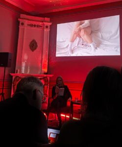 Kirjandusõhtu punases valguses: naine loeb mikrofoni ees raamatut, tema selja taga seinal on projektoripilt vereplekkidega valgel linal istuva inimese alakehast.