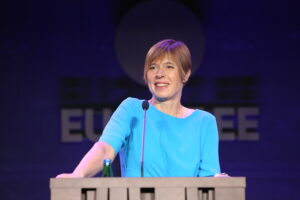 Kersti Kaljulaid seisab kõnepuldi taga ja naeratab, seljas lühikeste varrukatega helesinine kleit.