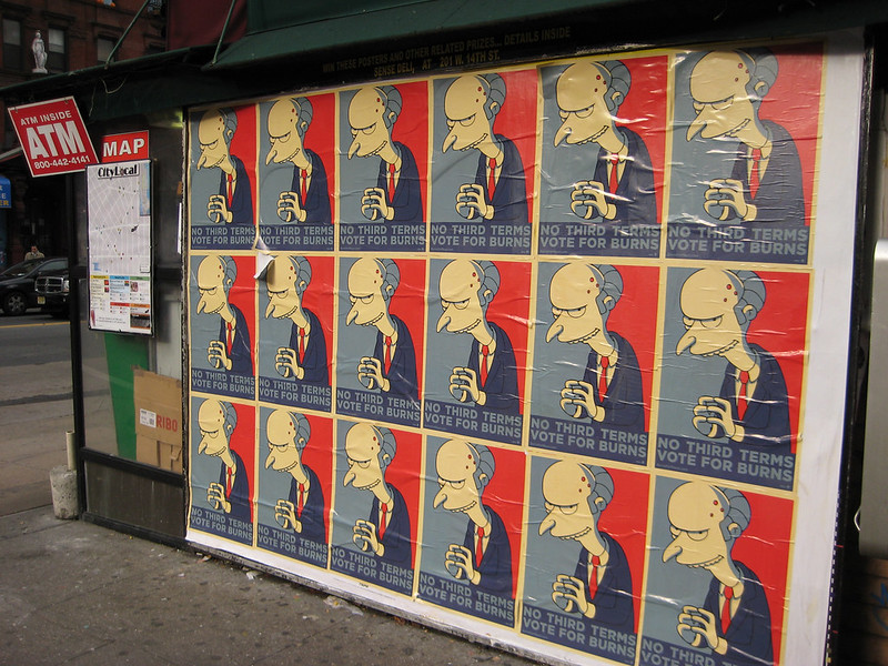 Seinale kleebitud plakatite kogum, mis kujutavad Montgomery Burnsi tegelast sarjast 'Simpsonid' propagandaplakatite stiilis. Plakatitel on punane taust ja tekst 'NO THIRD TERMS, VOTE FOR BURNS'.