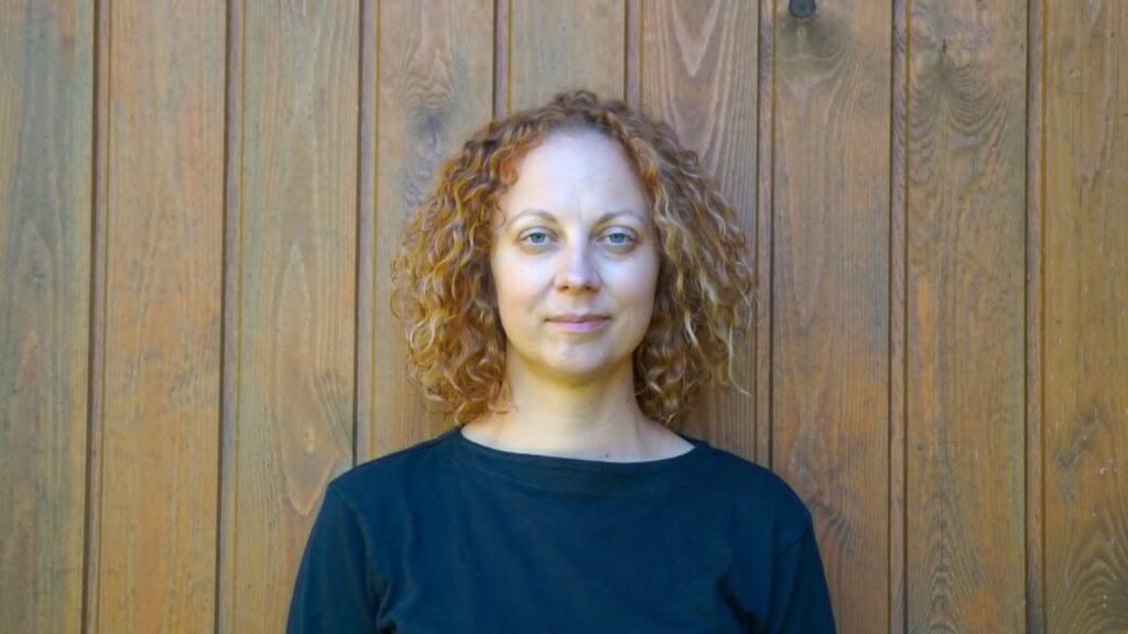 Portrait photograph of a woman with shoulder-length curly reddish-brown hair and blue eyes, wearing a dark blue boat-neck top, photographed against a light wooden wall with vertical panelling.