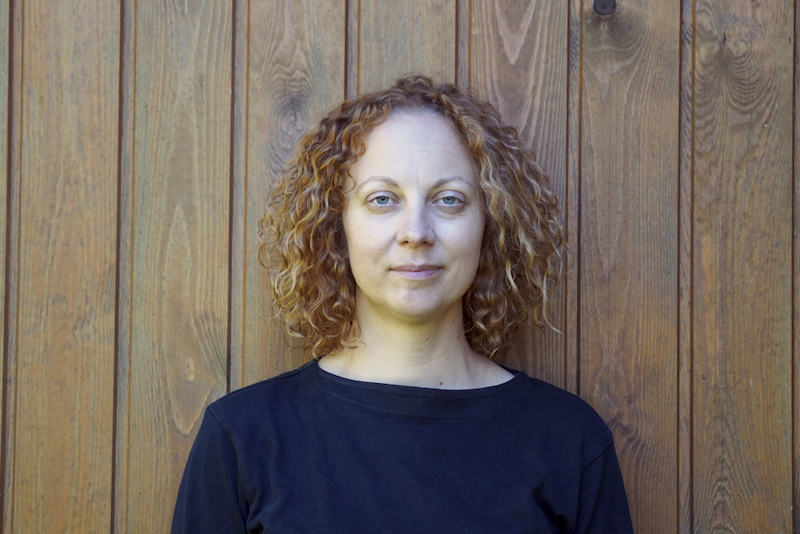 Portreefoto lokkis punakaspruunide juuste ja siniste silmadega naisest, kes kannab tumesinist paatkaelusega pluusi ja on pildistatud vertikaalsete paneelidega heleda puitseina taustal.