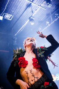 Drag king Dykeonaut performs on stage wearing a black blazer with elaborate shoulder decorations of greenery and red roses, torso decorated with drawn-on stitching, flat chest covered with red flowers. Photographed from below against blue stage lighting. Photo by Alecc Paalma.