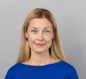 Portreefoto: heleda keskpikkade juustega naine sinises pluusis ja siniste kõrvarõngastega vaatab kaamerasse ning naeratab kergelt. Hall taust. 