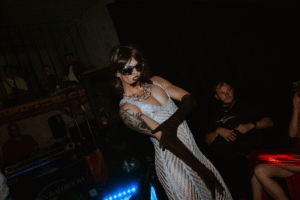 A drag performer wearing dark sunglasses and a glamorous silver beaded gown with a cutout design poses confidently with one hand on their hip, showing a visible arm tattoo. They wear an ornate crystal necklace and stand in a dimly lit venue with blue LED lights visible on equipment in the foreground and audience members seated in the shadowy background.