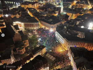 Õhuvaatefoto öisel ajal: tuhanded inimesed täidavad keskväljaku Läti vanalinnas. Rahvahulk on valgustatud mitmevärviliste tulede ja taskulampidega, mis loovad värvikirevaid mustreid. Ümberringi on ajaloolised hooned, mis on valgustatud soojade valgustega. Foto alumises nurgas on märgitud "POLICIJA".