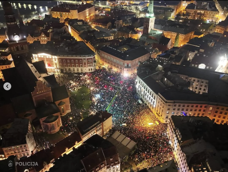 Õhuvaatefoto öisel ajal: tuhanded inimesed täidavad keskväljaku Läti vanalinnas. Rahvahulk on valgustatud mitmevärviliste tulede ja taskulampidega, mis loovad värvikirevaid mustreid. Ümberringi on ajaloolised hooned, mis on valgustatud soojade valgustega. Foto alumises nurgas on märgitud "POLICIJA".