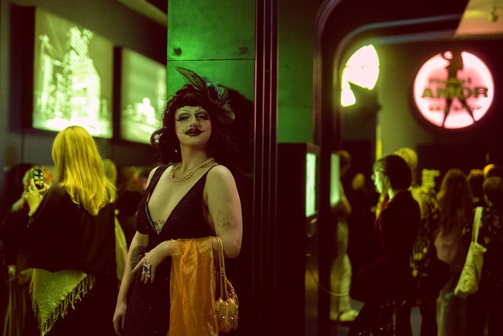 Drag performer Delphi Oraakel in a black and gold dress with pearl necklace and dramatic makeup, standing in a nightclub bathed in green lighting with crowd in background. Photo by Laila Kaasik.