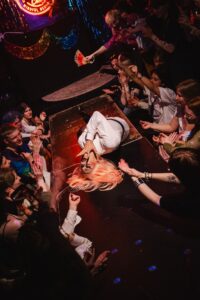 An overhead view of a drag performer in a white outfit with striking pink hair lying dramatically on the floor, surrounded by an enthusiastic crowd reaching out toward them with outstretched hands and phones. Colorful sequined decorations hang above, including gold and blue garlands, while warm stage lighting bathes the scene in amber and purple hues, creating an intimate nightclub atmosphere.