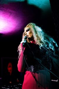 A performer with long, curly blonde hair and dramatic long eyelashes holds a microphone on stage, wearing a pink ribbed outfit with sparkly black embellishments. Dramatic purple and teal stage lighting creates an atmospheric glow against the dark background.