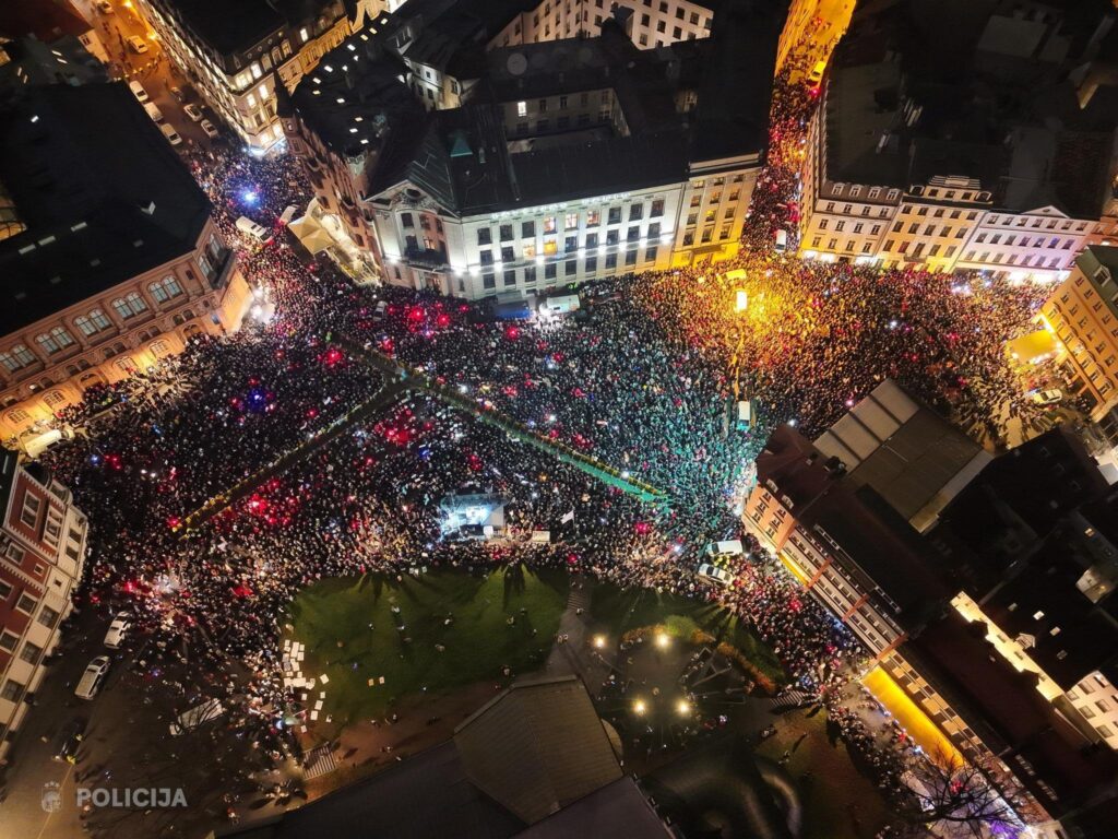 Aerial night photograph showing a massive nighttime protest filling a large city square and surrounding streets in Riga, with illuminated buildings around the perimeter and colorful lights visible throughout the crowd.