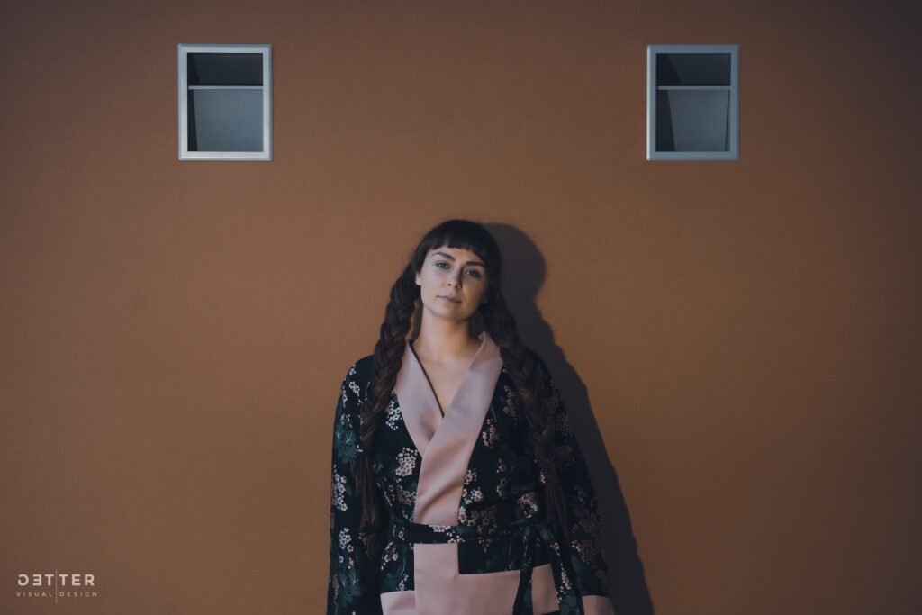 Foto autorist. Elise Rohtmets seisab pruunika seina ees, millel on kaks kõrgel asetsevat väikest akent. Tal on pikad pruunid palmikutesse põimitud juuksed ja lühike sirge tukk. Tema näoilme on tõsine. Tal on seljas lillemustriga tume kimonostiilis jakk roosade revääridega.