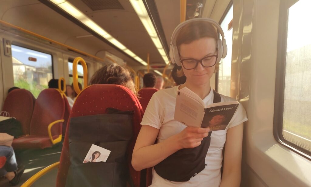 Sara Barbo istub rongis ja loeb raamatut. Tal on peas valged kõrvaklapid, seljas valge T-särk ja must vöökott. Käes hoiab ta Édouard Louis'i raamatut.